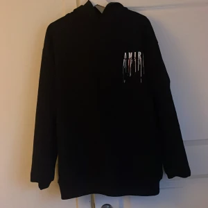 Svart AMIRI hoodie med färgstänk - Svart hoodie från AMIRI med coolt färgstänksmönster och stor logga på ryggen. Tröjan har huva och långa ärmar, samt tryck på bröstet och ryggen i vitt, blått och orange. Perfekt för dig som gillar streetwear och vill sticka ut.