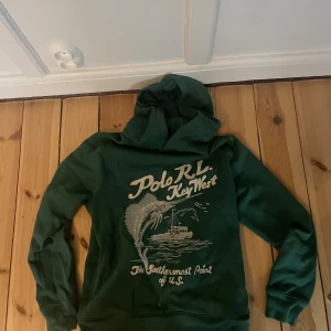 Grön hoodie Polo Ralph Lauren L - Snygg grön hoodie från Polo Ralph Lauren i storlek L med tryck på framsidan som visar fisk och båt samt texten 'Polo RL Key West'. Tröjan har huva och långa ärmar, tillverkad i mjuk bomull. Perfekt för chill dagar och streetwear.