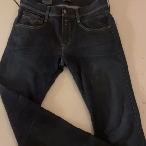 Replay Anbass mörkblå jeans 29/32 - Snygga mörkblå Replay Anbass jeans med klassisk femficksdesign och kontrastsömmar. Modellen har raka ben och normal passform, tillverkade i slitstarkt jeanstyg. Bakfickorna har stilrena broderade detaljer som ger extra edge till looken.