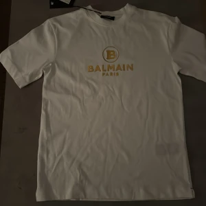 Vit Balmain t-shirt med guldtryck - Vit t-shirt från Balmain med klassiskt guldtryck på bröstet där det står 'BALMAIN PARIS' och en stor B-logga. T-shirten har rund hals och korta ärmar, tillverkad i mjuk bomull för en clean och lyxig vibe. Perfekt för dig som gillar exklusiva märken. Äkta med kvitto 