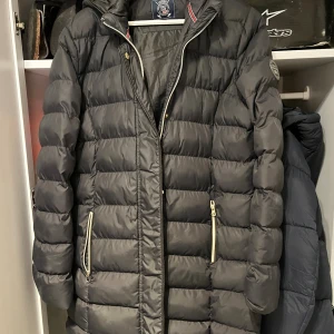 Svart lång pufferjacka Vinson Polo Club - Säljer en svart lång pufferjacka från Vinson Polo Club med huva och snygga vita dragkedjor på fickorna. Jackan har quiltad design och ett stilrent märke på ärmen. Perfekt för kalla dagar och har en modern look som passar till streetwear.