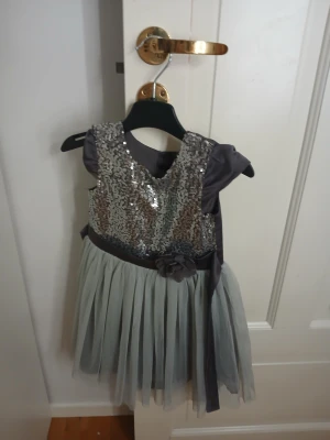 Grå kort klänning med paljetter och rosett - Superfin grå kort klänning med glittrig paljettöverdel och puffärmar. Kjolen är i mjuk tyll och midjan har ett satinband med en dekorativ rosett. Klänningen har knappar i ryggen och passar perfekt för speciella tillfällen.