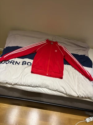 Röd Adidas vindjacka med vita ränder, - Säljer en helt oanvänd röd Adidas vindjacka med klassiska vita ränder längs ärmarna och Adidas-logga på bröstet. Jackan har dragkedja framtill, två fickor med dragkedja och ribbade muddar. Perfekt för dig som gillar sportig och retro stil.