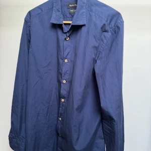 Blå skjorta från Massimo Dutti M - Snygg blå skjorta från Massimo Dutti i storlek M. Skjortan har klassisk krage, långa ärmar och träfärgade knappar framtill. Tillverkad av bomull, perfekt för dig som gillar stilrena och enkla plagg med bra passform.