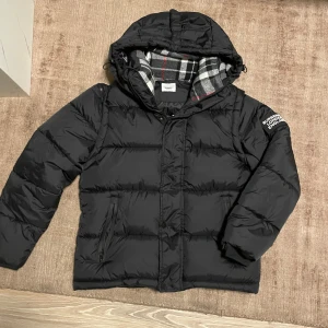 Burberry jacka  - Svart pufferjacka från Burberry med avtagbara ärmar så du kan bära den som väst. Jackan har huva med snörning och ett rutigt foder i grått, vitt och rött. Logotypen 'BURBERRY LONDON ENGLAND' är tryckt på vänster ärm. Dragkedja och fickor framtill. Stl S jätte bra skick och tags medföljer. 