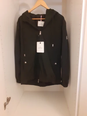 Svart vindjacka från Moncler - Svart vindjacka från Moncler med huva och dragkedja framtill. Jackan har Moncler-logga på ärmen och snygga detaljer i rött, vitt och blått på huvan. Tillverkad i lätt polyester, perfekt för blåsiga dagar. Fickor med tryckknappar och justerbar snörning vid huvan.