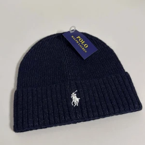 Mörkblå ribbad mössa Polo Ralph Lauren - Snygg mörkblå ribbstickad mössa från Polo Ralph Lauren med klassisk broderad logga i vitt framtill. Tillverkad i en mjuk blandning av bomull och polyester, perfekt för kyliga dagar. Enkel och stilren design som lyfter din look.