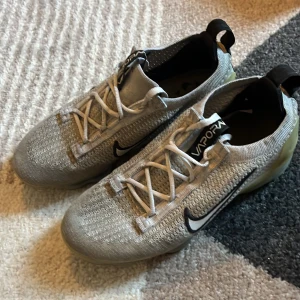 Nike Vapormax grå sneakers - Nike Vapormax sneakers i ljusgrå stickad mesh med vita snören och svart swoosh på sidan. Sulan är transparent med inslag av beige och svarta prickar, vilket ger en unik look. Skorna har platt sula och dragflik bak för enkel påtagning. Perfekta för dig som gillar sportig stil.