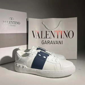 Valentino Garavani vita sneakers - Snygga vita Valentino Garavani sneakers med bred marinblå rem över sidan och coola nitar på hälen. Skorna har rund tå, platt sula och snörning framtill. Tillverkade i skinn för en lyxig känsla och stilren look. Använda 1 gång. OBS!!! Ingen låda eller påse på bilden följes med bara skorna!