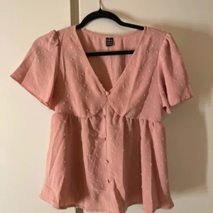 Rosa blus med v-ringning från SHEIN - Supersöt ljusrosa blus från SHEIN med v-ringning, korta ärmar och knappar framtill. Blusen har ett diskret broderat prickmönster och lätt rynkad midja för en flowy look. Perfekt för dig som gillar romantiska och feminina detaljer.