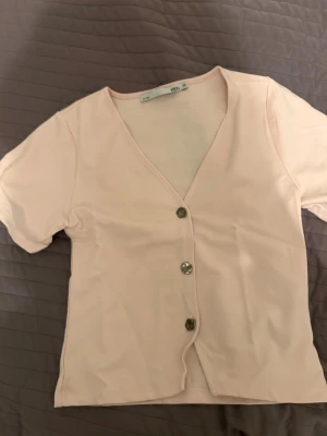 Ljusrosa blus med v-ringning och knappar - Söt ljusrosa blus med v-ringning och tre dekorativa knappar framtill. Kortärmad och i mjukt material, ej använd!