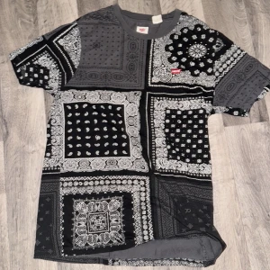 Svart och grå bandana t-shirt Levi's - Säljer en unik t-shirt från Levi's i storlek S med svart och grå patchwork-bandana mönster. T-shirten har korta ärmar, rund hals och ett litet rött Levi's-märke på bröstet. Materialet är mjuk bomull och passformen är normal.