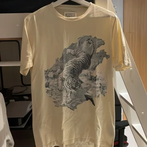 Beige t-shirt med tigertryck Maison Margiela - Snygg kräm vit t-shirt från Maison Margiela med ett stort, detaljerat tigertryck på framsidan. Modellen är klassisk med rund hals och korta ärmar. Materialet är mjuk bomull som känns skönt mot huden. Perfekt för dig som vill sticka ut med ett coolt motiv. Strlk 50/m-l