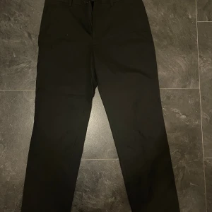 Svarta kostymbyxor med raka ben - Snygga svarta kostymbyxor med klassisk passform och raka ben. Byxorna har hällor för bälte, diskreta bakfickor med knapp och stängs med dragkedja och knapp framtill. Perfekta för en clean och stilren look.