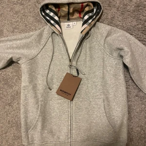 Grå hoodie från Burberry med dragkedja - Snygg grå hoodie från Burberry med klassiskt rutigt foder i huvan och dragkedja framtill. Tröjan har snörning i huvan, långa ärmar och två fickor. Perfekt för dig som vill ha en stilren och lyxig look.