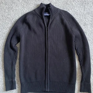 Svart stickad kofta från Marwin & Friends - Svart stickad kofta från Marwin & Friends med hel dragkedja framtill och ribbade muddar. Hög krage och långärmad modell, perfekt för lager-på-lager. Enkel och stilren design som funkar till det mesta. Storlek S.