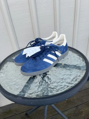 Adidas Spezial blå sneakers mocka - Helt nya, endast provade. Adidas godkännde ej retur. Nypris 1300Säljer ett par Adidas Gazelle sneakers i blå mocka med vita detaljer och klassiska tre ränder på sidan. Skorna har snörning, grå platt sula och vit insida med Adidas-logga. Stilren och retro design som passar perfekt till streetwear. Som även har ett par extra vita snören. 