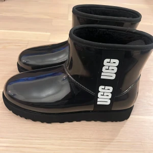 Svarta UGG boots med glansig yta - Säljer ett par svarta UGG boots med glansig syntetisk yta och bred UGG-logga på sidan. Insidan är fodrad med mjuk päls för extra komfort och värme. Rund tå och platt sula gör dem sköna att bära. Perfekta för kalla dagar och har en modern look. Som ny. Använda endast ett par ggr. Har vanligtvis strl 38 i de flesta skor men i just Uggs har jag strl 39. 