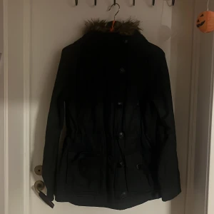 Svart parkajacka med fuskpäls från H&M - Svart parkajacka från H&M L.O.G.G. med snygg fuskpäls på huvan. Jackan har knappar framtill, två stora fickor och dragsko i midjan för en schysst passform. Perfekt för kalla dagar med sin långa modell och klassiska look.