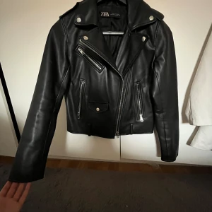 Svart skinnjacka från Zara - Snygg svart skinnjacka från Zara. Jackan har flera silvriga dragkedjor, knappar och fickor framtill. bra skick förutom slitningar i kragen (sista bilden), däremot inget som syns då det är innanför samt i nacken. nypris 500kr 