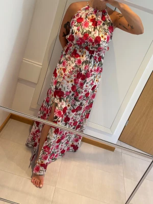 Blommig röd långklänning från Gina Tricot - Supersnygg långklänning från Gina Tricot med blommigt mönster i rött, rosa och gult på vit botten. Klänningen har halterneck, elastisk midja och en slits framtill som ger extra flow. Drömmig maxiklänning med blommigt mönster, slits, halterneck och vit underklänning.