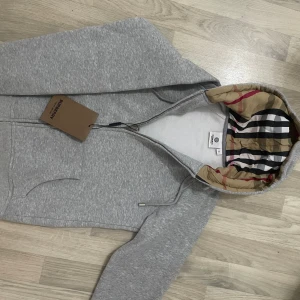 Grå Burberry hoodie med klassiskt foder - Grå hoodie från Burberry med dragkedja och huva. Insidan av huvan har det ikoniska rutiga Burberry-mönstret i beige, svart, rött och vitt. Tröjan är i mjuk bomullsmix och har broderad logga på bröstet. Perfekt för en stilren och lyxig vardagslook.