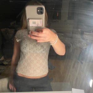 Vit spetsblus med kort ärm - Säljer en söt vit blus i spets med korta ärmar och lätt volangkant vid hals och ärmslut. Blusen är genombruten och har ett fint mönster i tyget, perfekt att styla lager på lager. Modellen är figurnära och croppad.