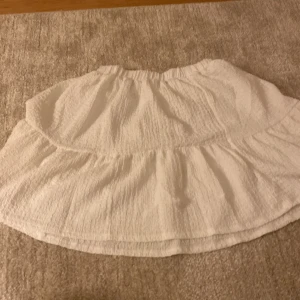 Vit volangkjol med resårmidja - Säljer en vit kortkjol med volang och resår i midjan. Kjolen har en lätt skrynklig struktur och är gjord i ett mjukt bomullstyg. Perfekt för dig som gillar en luftig och chill stil. Passar dig som vill ha en enkel men söt kjol med volangdetalj.