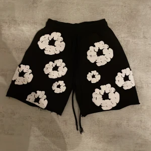 Svarta Denim Tears shorts  - Svarta shorts från Denim Tears med vita blommiga tryck över hela plagget. De har råa kanter nertill och snörning i midjan för en avslappnad vibe. Perfekta för sommaren och sticker ut med sitt unika mönster. Skick 9/10 hade varigt 10/10 fast e lite slitna på snörena. Skriv för mer information/ bilder, mvh John