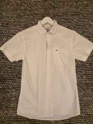 Vit kortärmad skjorta Tommy Hilfiger - Vit skjorta från Tommy Hilfigur i storlek S med klassisk krage och korta ärmar. Regular fit med bröstficka och broderad logotyp. Skjortan har knappar framtill och är gjord i mjuk bomull, perfekt för en clean look