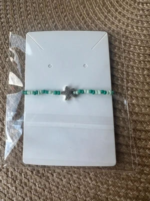 Turkost pärlarmband med sjöstjärna - Snyggt hemmagjort armband med små turkosa och vita pärlor, dekorerat med en silverfärgad sjöstjärna i mitten. Perfekt accessoar för dig som gillar strandvibbar och detaljer som sticker ut. Armbandet är enkelt och stilrent, passar till många olika looks.
