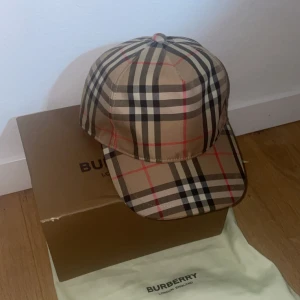 Burberry keps i klassiskt rutmönster - Säljer en snygg keps från Burberry med det ikoniska beigea rutmönstret och röda detaljer. Kepsen har böjd skärm perfekt för dig som vill ha en stilren accessoar med lyxig känsla.
