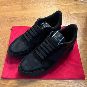 Valentino Garavani svarta sneakers - Svarta Valentino Garavani sneakers med coolt camouflage-mönster och snygga detaljer i mocka och skinn. Platt sula med nitar på sidan och rund tå. Skorna har snörning och en sportig vibe, perfekt för dig som gillar exklusiva märken och streetstyle.