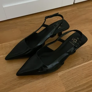 Svarta pumps med klack från VOX - Snygga svarta pumps från VOX i veganskt material med spetsig tå och låg klack. Skorna har en elegant rem med spänne runt hälen och en stilren, minimalistisk look. Perfekt för dig som gillar trendiga och klassiska detaljer.