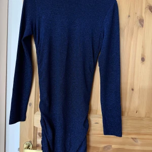 Mörkblå glittrig långärmad klänning - Snygg mörkblå festklänning med glittrigt tyg och lång ärm. Modellen är figurnära med rynkade detaljer längs sidorna och har en kort längd. 