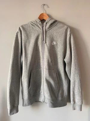 Grå hoodie från Nike med dragkedja - Snygg grå hoodie från Nike i storlek M. Tröjan har huva med snörning, två fickor fram och hel dragkedja. Tillverkad i mjuk bomullsblandning och har Nike-logga på bröstet. Perfekt för chill eller träning.