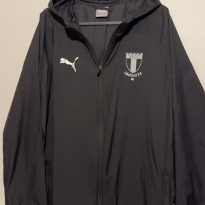 MALMÖ FF - Allwetterjacka PUMA - Svart vindjacka från Puma med Malmö FF-logga på bröstet och vit Puma-logga. Jackan har huva, hel dragkedja och är tillverkad i lätt polyester. Perfekt för dig som vill representera MFF och hålla dig skyddad mot vinden. (Knappast använd)