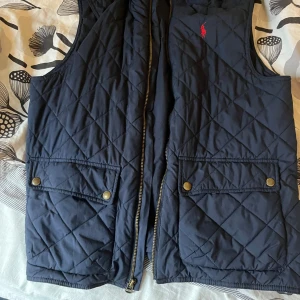 Polo Ralph Lauren väst  - Snygg quiltad dunväst från Polo Ralph Lauren i marinblått med guldfärgade tryckknappar och dragkedja. Västen har två stora fickor framtill och klassisk röd Polo-logga på bröstet. Perfekt för lager-på-lager och chill stil.