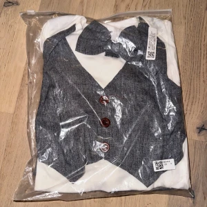 Festlig babykostym Grå kostymset med fluga för barn - Snyggt tvådelat kostymset för barn med vit långärmad body och fastsydd grå väst med bruna knappar och matchande grå byxor. Setet har även en söt fluga i samma tyg som västen och byxorna. Perfekt för speciella tillfällen när man vill vara extra uppklädd.Festlig babykostym med väst och fluga. Inkluderar även ett par byxor. Perfekt för speciella tillfällen.