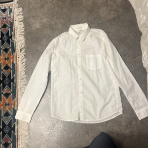 Vit skjorta från H&M Easy Iron - Klassisk vit skjorta från H&M Easy Iron med lång ärm, bröstficka och knappar hela vägen framtill. Skjortan är i bomull och har en stilren, tidlös look som passar till många olika outfits. Perfekt för dig som gillar enkel och clean stil.