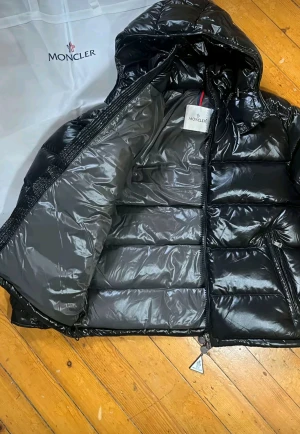 Svart Moncler pufferjacka med huva - Säljer en svart pufferjacka från Moncler med glansig finish och huva. Jackan har Moncler-logga på ärmen, dragkedja framtill och två sidofickor med dragkedja. Perfekt för kalla dagar och har en riktigt schysst streetstil.