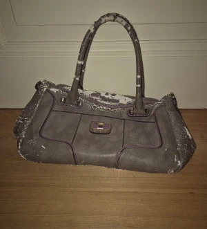 Guess handväska i grått med lila detaljer - Fin handväska från Guess i grått skinnimitation med lila detaljer och spänne framtill. Väskan har reptilmönstrad yta och insida med leopardmönster. Två handtag och avtagbar lila axelrem. Finns 3 små fack och ett stort utrymme inuti väskan. Det flagnar lite på handtagen och på sidorna men annars i fint skick💓