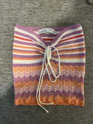 Randig croppad topp från Bershka - Superfin croppad topp från Bershka i storlek S. Toppen har ett färgglatt randigt mönster i lila, orange, vit och röd med snygg snörning framtill. Materialet är ribbad och stretchigt, perfekt för dig som gillar att sticka ut med coola färger och detaljer.