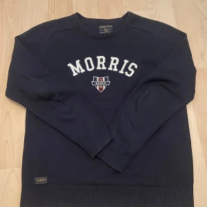 Mörkblå stickad tröja från Morris - Snygg mörkblå stickad tröja från Morris med broderad logga och text framtill. Tröjan är i 100% bomull, har rund halsringning. Perfekt för dig som gillar klassisk stil. Storlek L!