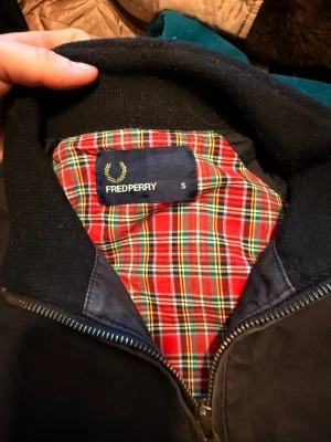 Svart bomberjacka från Fred Perry - Svart bomberjacka från Fred Perry med klassisk gul logga på bröstet. Jackan har dragkedja, ribbade muddar och fickor med bruna knappar. Insidan har ett snyggt röd-rutigt foder  som ger extra stilpoäng. Perfekt för dig som gillar street och retrovibe.