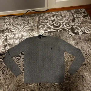 Grå kabelstickad Raulph Lauren tröja - Säljer en grå stickad tröja från Ralph Lauren. Perfekt för dig som gillar stilren och tidlös design.