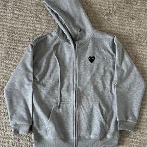 Grå hoodie från Comme des Garçons Play - Grå hoodie med dragkedja från Comme des Garçons Play. Ikoniskt svart hjärta med ögon på bröstet och stort tryck på ryggen. Huva med snörning och två fickor framtill. Tillverkad i mjuk bomull för en chill och stilren look.