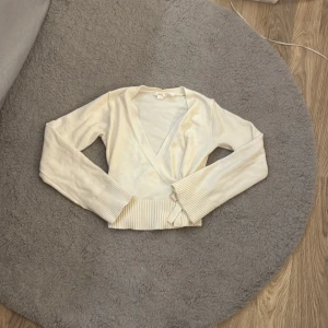 Beige omlottkofta med knytband - Superfin beige omlottkofta med långa ärmar och bred ribbad mudd. Knytband i midjan för en snygg och feminin look. Perfekt att slänga över en topp för extra stilpoäng.