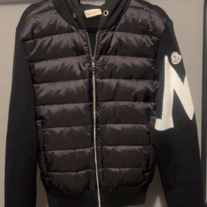 Svart pufferjacka från Moncler - Svart pufferjacka från Moncler med stickade ärmar och stor vit M-detalj på ena ärmen. Jackan har huva, dragkedja framtill och quiltad front i syntetmaterial. Perfekt för dig som vill ha en cool och sportig look med premiumkänsla.