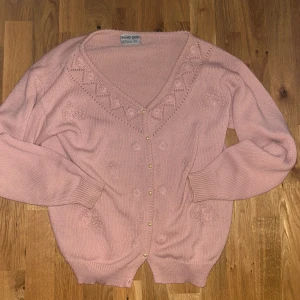 Rosa kofta med pärlknappar från Knit Set - Supersöt ljusrosa kofta med v-ringning och pärlknappar från Knit Set. Stickad med blommiga detaljer och mönster runt halsen. Långärmad och mjuk, perfekt för att ge din outfit en gullig touch. Ribbad vid ärmslut och nederkant för extra komfort.
